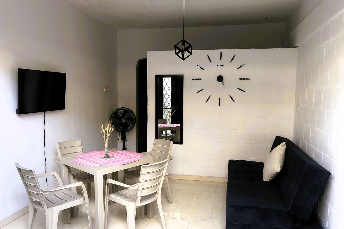 Un coin repas comprenant une table, des chaises, une horloge, un téléviseur et un canapé noir.