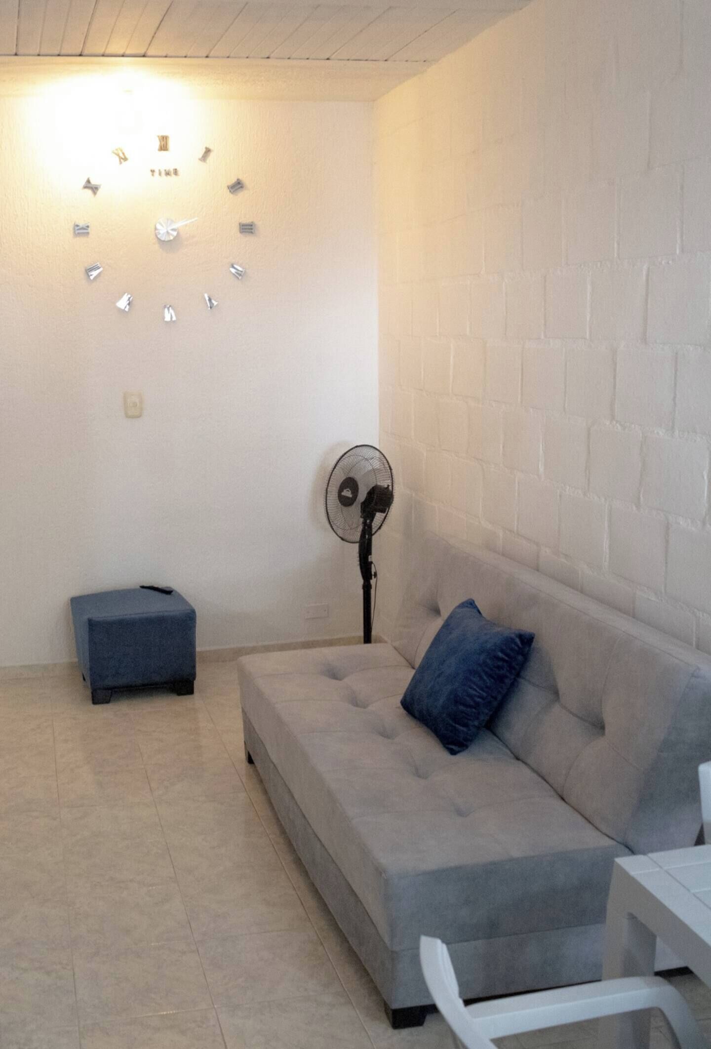 Una habitación con un sofá gris, un otoman azul pequeño, una mesa blanca, un reloj de pared y un ventilador de pie.