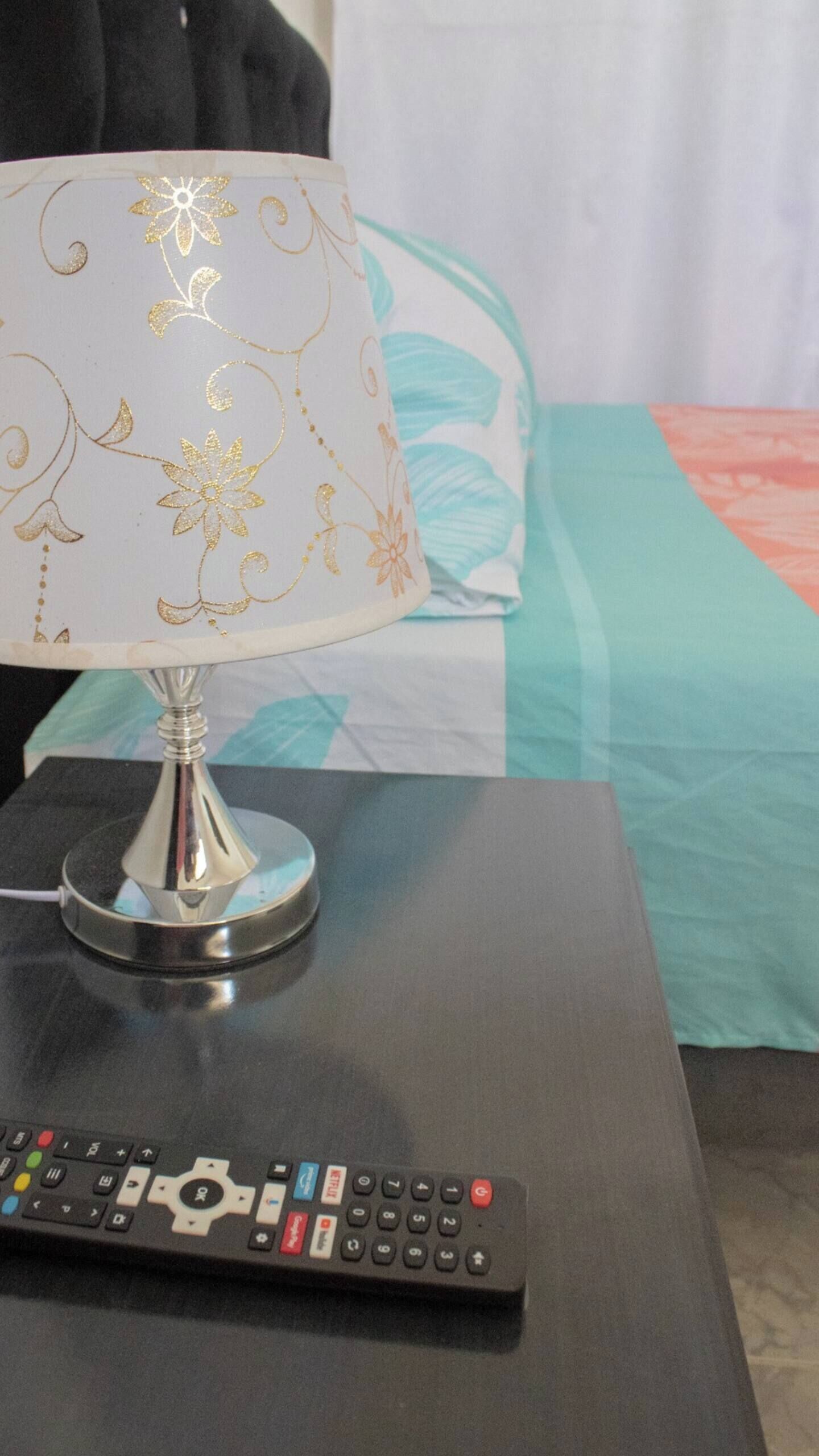 Een bed met een blauwgroene en oranje sprei, een witte lamp met gouden bloemmotieven, en een afstandsbediening op een zwart nachtkastje.