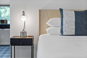 A neatly made bed with white and blue bedding, a bedside lamp, and a small wooden nightstand with a drawer.