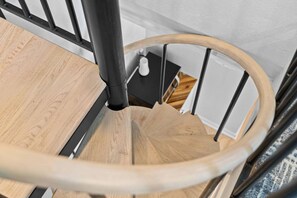 A wooden spiral staircase with a black metal railing and a white wall in the background.