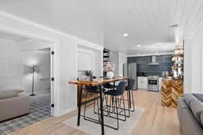 Eine moderne Küche mit Kochinsel, ein Essbereich mit Holztisch und Barhocker sowie ein Wohnzimmer mit Sofa und Stehlampe.