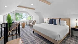 A bedroom with a large bed, a wooden headboard, a window with a view of greenery, a small table with a lamp, and a staircase with a railing.