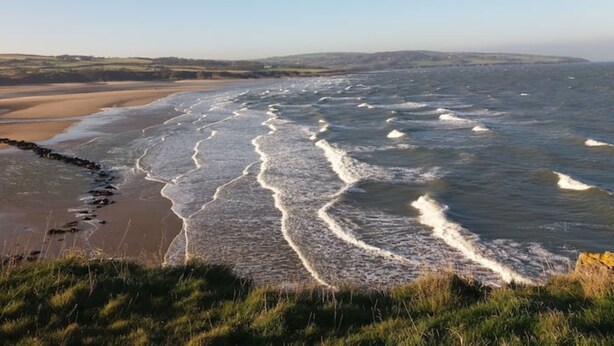 Un paisaje costero con olas rompiendo en la orilla, una playa de arena y zonas cubiertas de hierba.