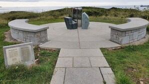 Una estructura de piedra circular con una placa, rodeada de césped y un cuerpo de agua al fondo.