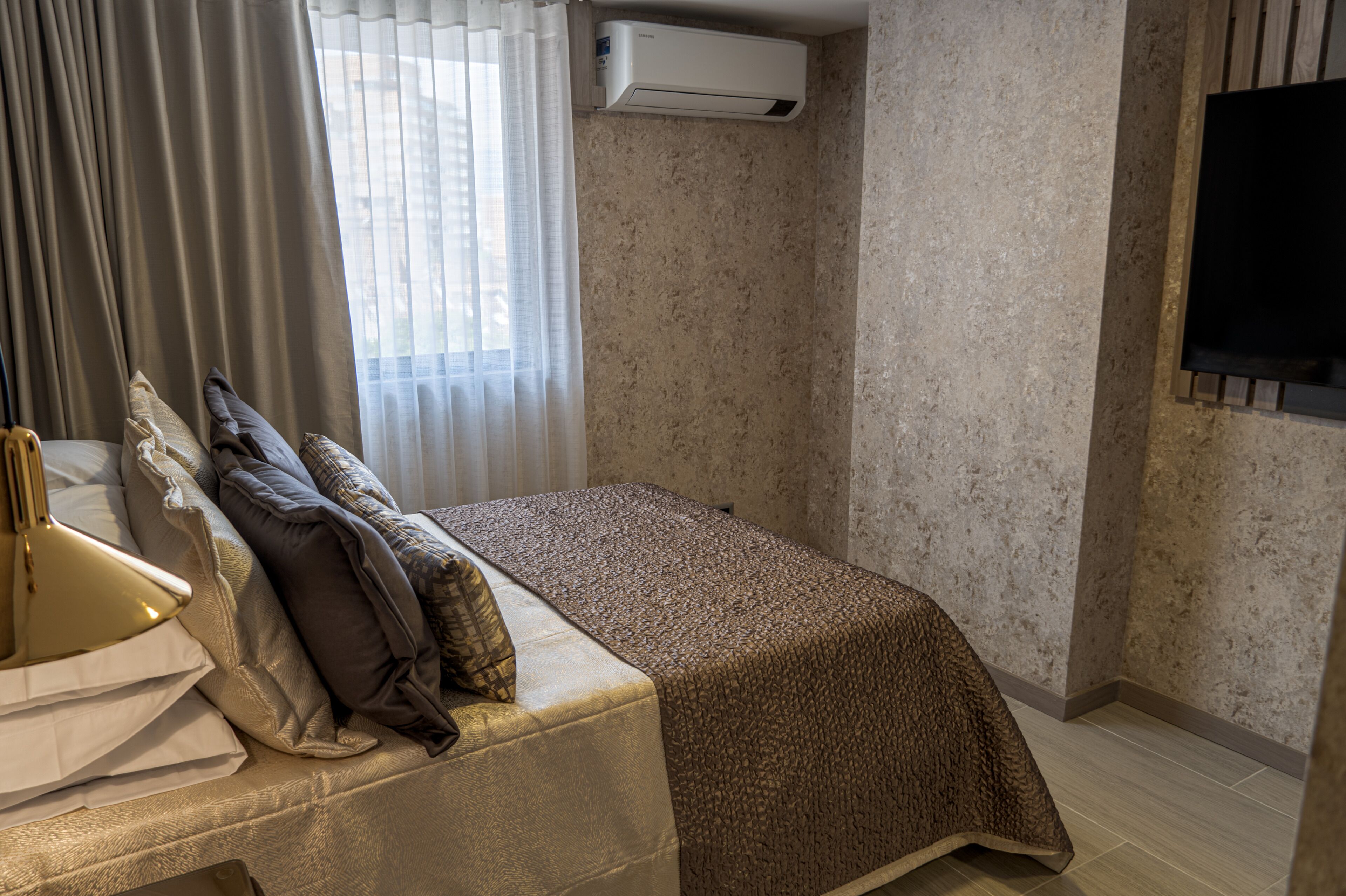 Una habitación de hotel con cama, escritorio con lámpara, ventana con cortinas, unidad de aire acondicionado y televisor montado en la pared.