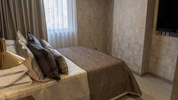 Una habitación de hotel con cama, escritorio con lámpara, ventana con cortinas, unidad de aire acondicionado y televisor montado en la pared.