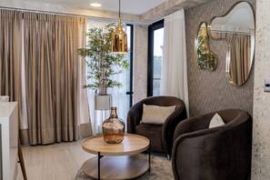 A modern living room with a round wooden table, two dark armchairs, a hanging light fixture, a large mirror, and a potted plant. - Luxury Suites Prana By Cadissa (Medellín)