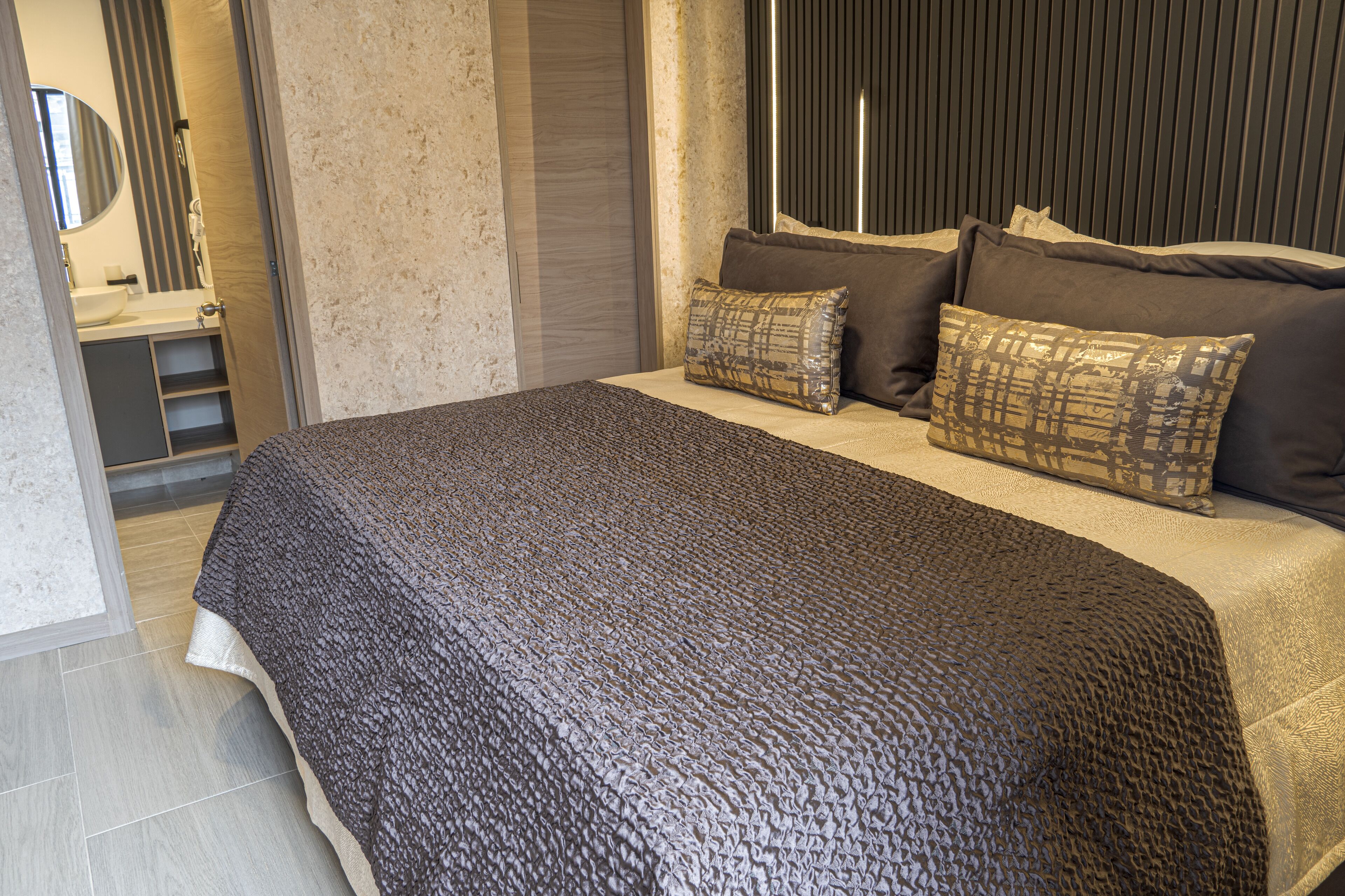 Een moderne slaapkamer met een groot bed, meerdere kussens, een nachtkastje en een badkamer die door een open deur zichtbaar is.