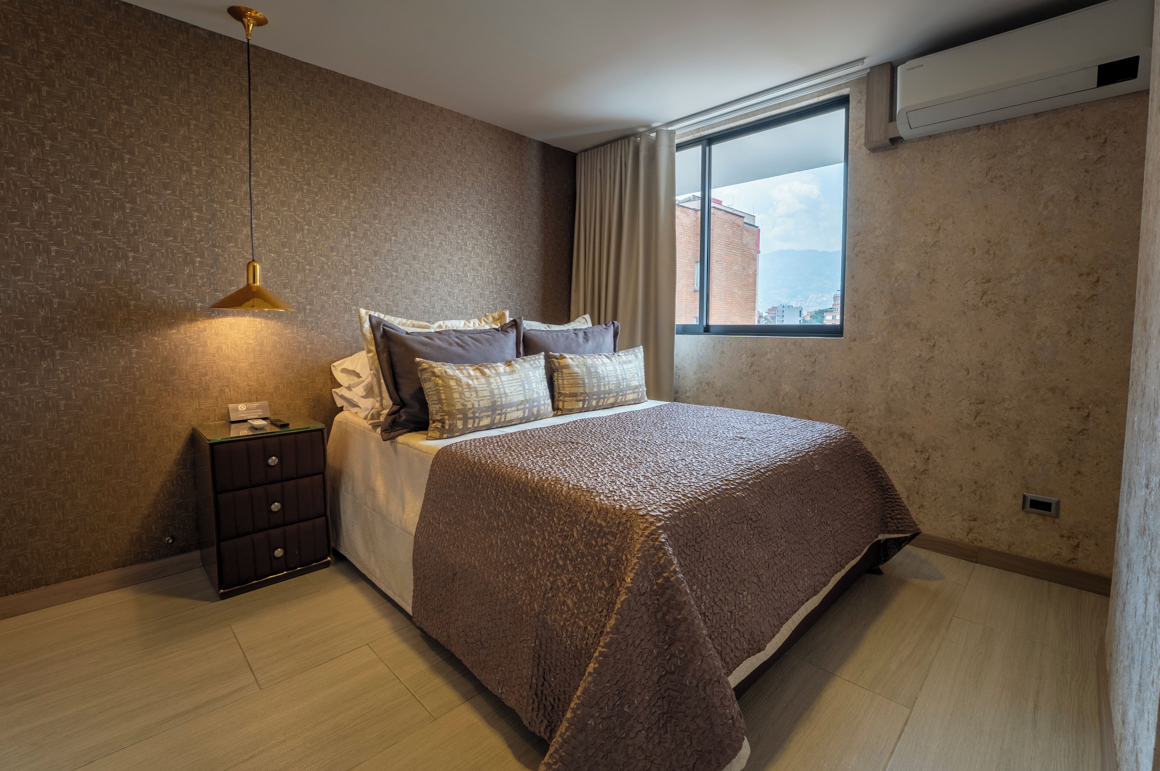 A modern bedroom with a large bed, a bedside table, a lamp, and a window with a view of buildings.