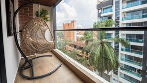 Un balcon avec un fauteuil en osier, une vue sur les immeubles voisins et un palmier.
