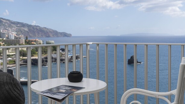 A balcony with a white table, a black vase, and a magazine. The view includes a body of water and distant land.