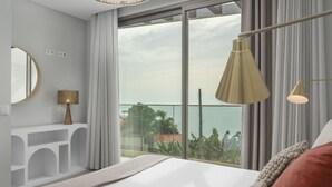 Ein modernes Hotelzimmer mit einem großen Bett, einem Nachttisch, einer Lampe und Blick auf das Meer durch raumhohe Fenster.