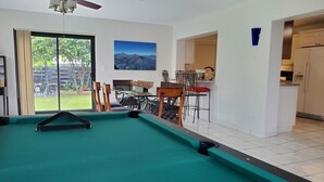 A pool table in a room with a ceiling fan, a dining area with chairs and a table, and a kitchen visible through an open doorway. - Cozy family home in quiet area, nice garden views. (Miami)