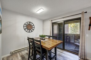 Un coin repas avec une table et des chaises en bois, une horloge au mur et une porte-fenêtre coulissante donnant sur une terrasse extérieure.