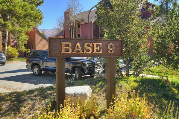 Una señal con la leyenda 'BASE 9' en una zona residencial con casas, vehículos estacionados y vegetación.