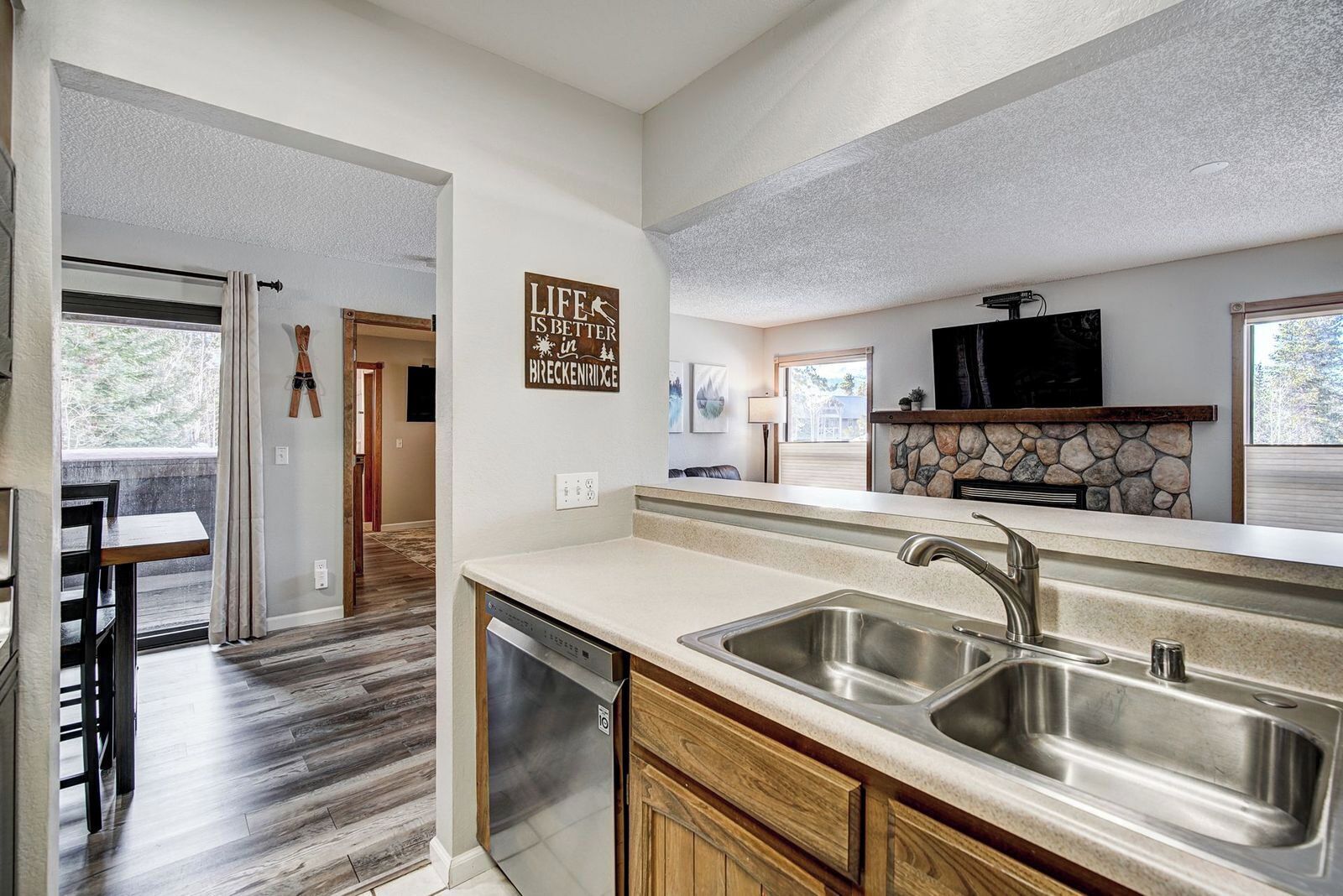 A modern kitchen with a double sink, wooden cabinets, a stone fireplace, and a dining area with a view.
