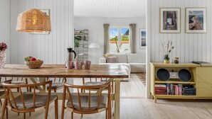 Un salon spacieux avec une table à manger en bois, des chaises rembourrées, un luminaire suspendu en osier, un canapé blanc, une bibliothèque et des tableaux encadrés accrochés au mur.