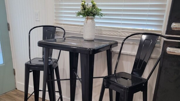 A kitchen with a black metal table and chairs, a white vase with green plants, and a window with blinds.
