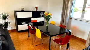 Un salon moderne avec une table à manger noire, des chaises rouges et jaunes, un meuble de rangement noir et blanc pour les appareils électroniques, et une plante en pot.