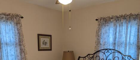 A bedroom with a bed, a ceiling fan, a nightstand with a lamp, and curtains.