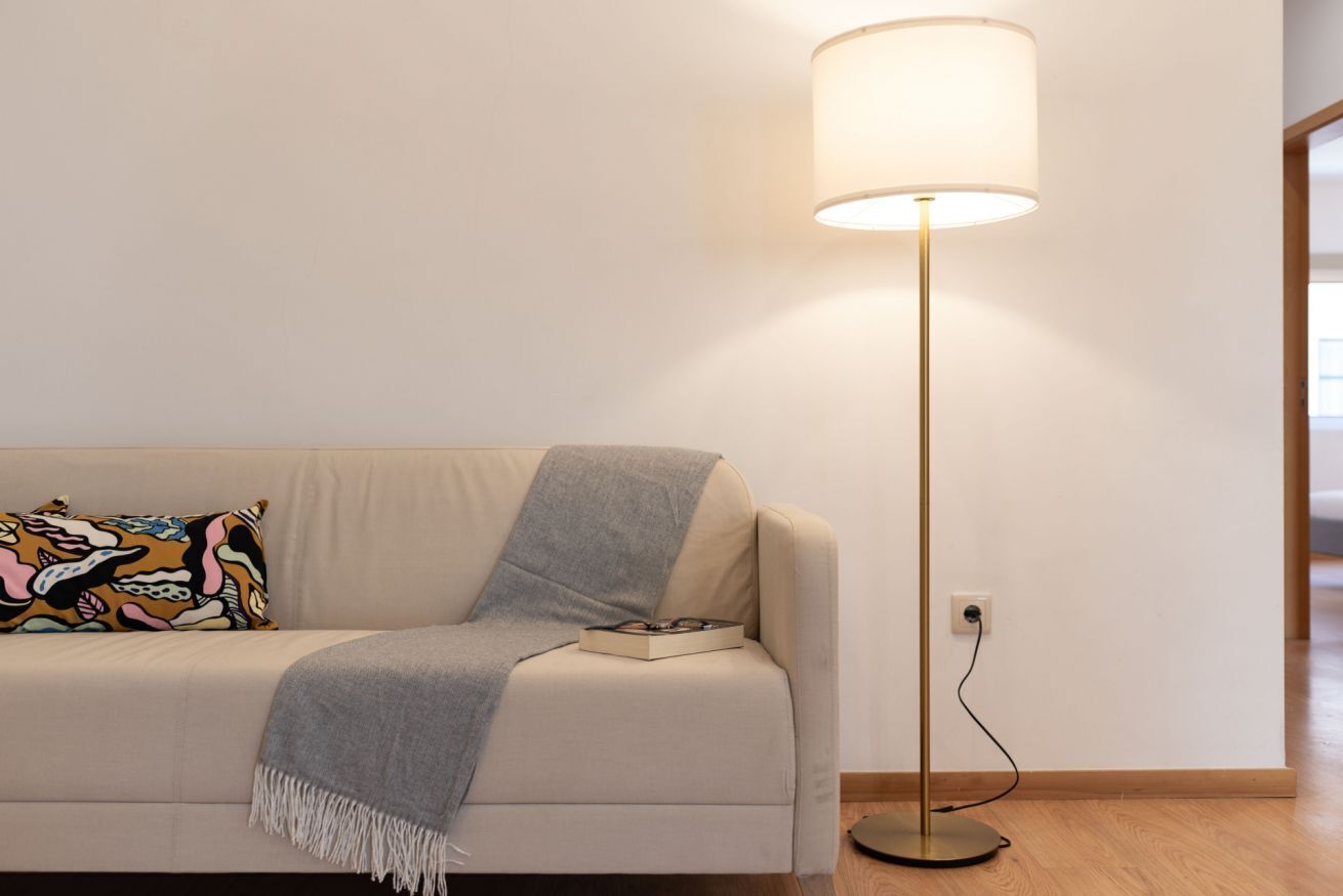 Ein modernes Wohnzimmer mit einem beigen Sofa, einem farbenfrohen Kissen, einer grauen Decke und einer Stehlampe mit weißem Lampenschirm.