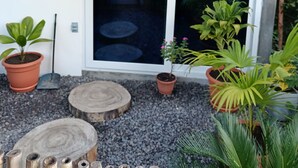 Une construction à toit de chaume dotée d’une porte coulissante en verre, entourée de plantes en pot et d’une bûche de bois.