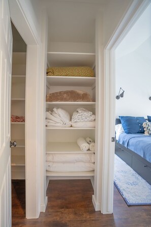 Ein begehbarer Kleiderschrank mit mehreren Einlegeböden für gefaltete Kleidung, ein Bett mit blauer Bettdecke und ein grauer Kopfteil.