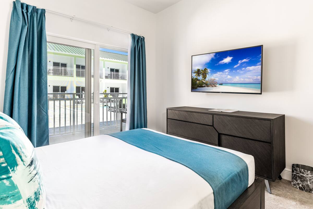 A hotel room with a bed, a dark wooden dresser, a television on the wall, and a balcony with a view of a building and palm trees.