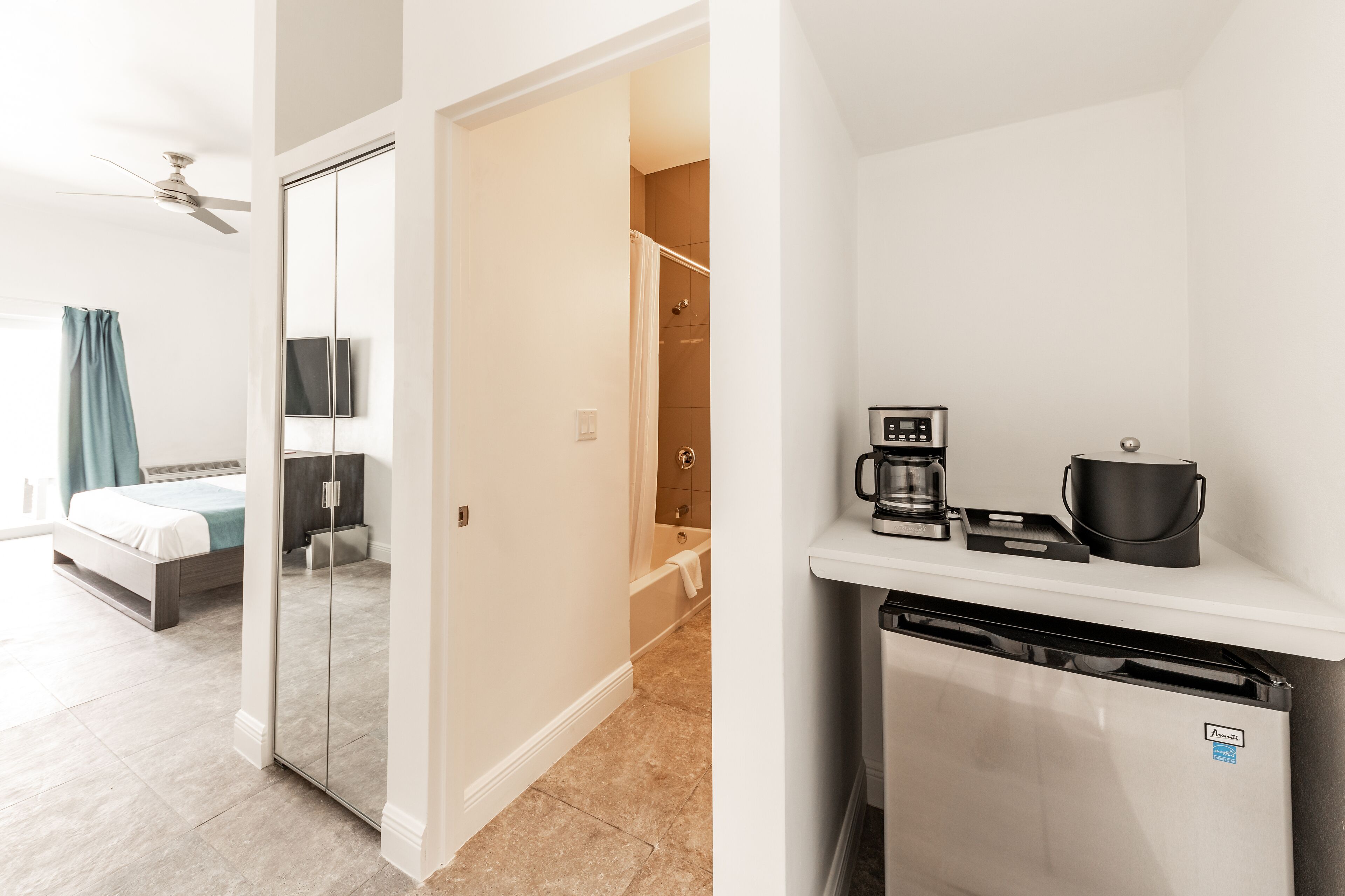 A modern hotel room with a bed, a bathroom with a shower and bathtub, and a kitchenette with a coffee maker and a small refrigerator.