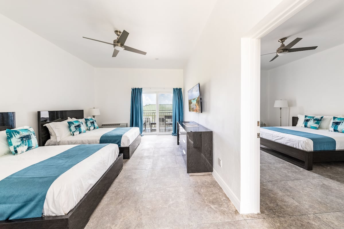A modern hotel room with two beds, a desk, and a balcony with blue curtains.
