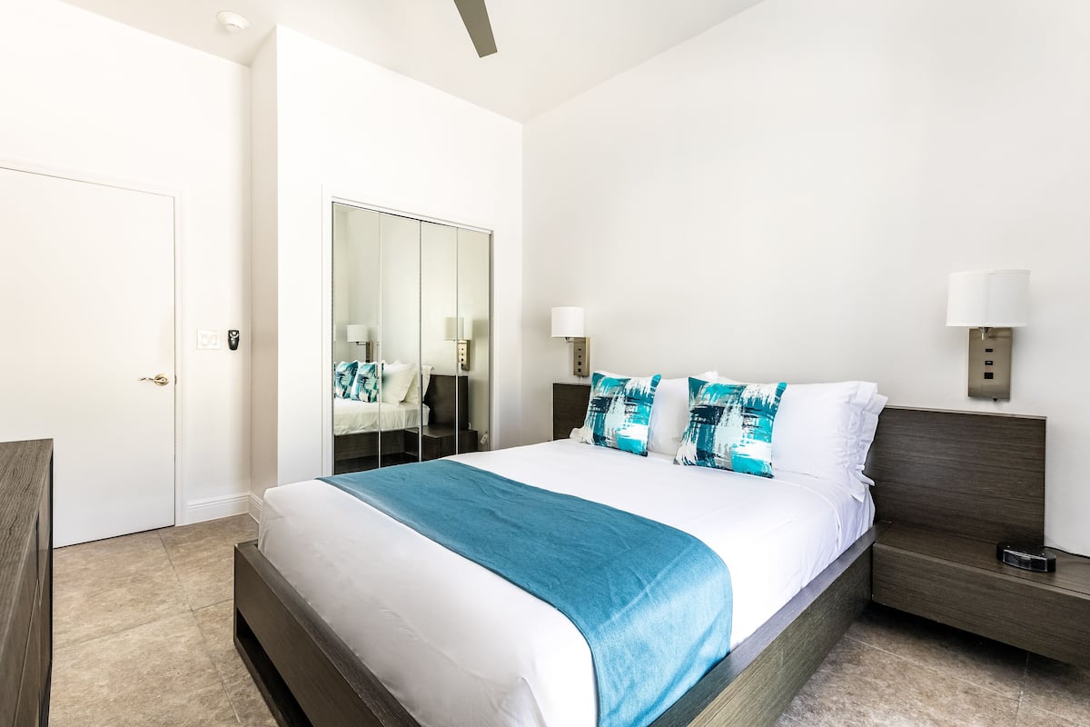 A modern hotel room with a large bed, a bedside table, a nightstand, a mirror, and a wardrobe.