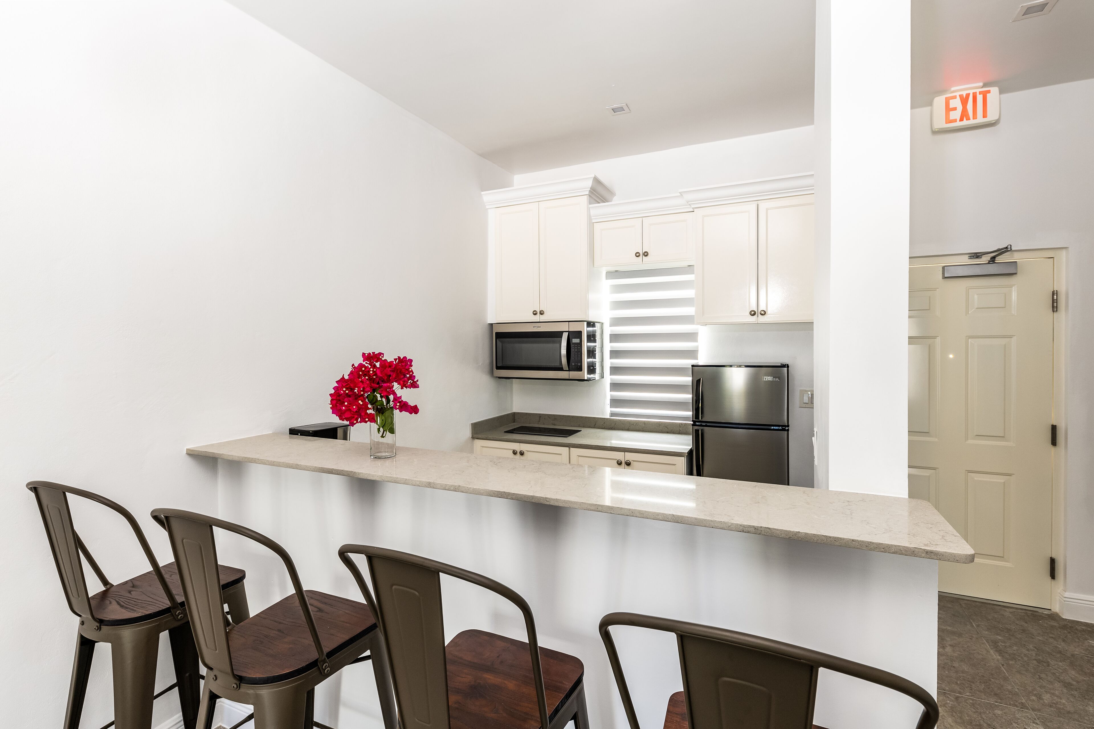 Una cocina moderna con armarios blancos, electrodomésticos de acero inoxidable y encimera de mármol. Hay cuatro taburetes de bar y un florero con flores rojas sobre la encimera.