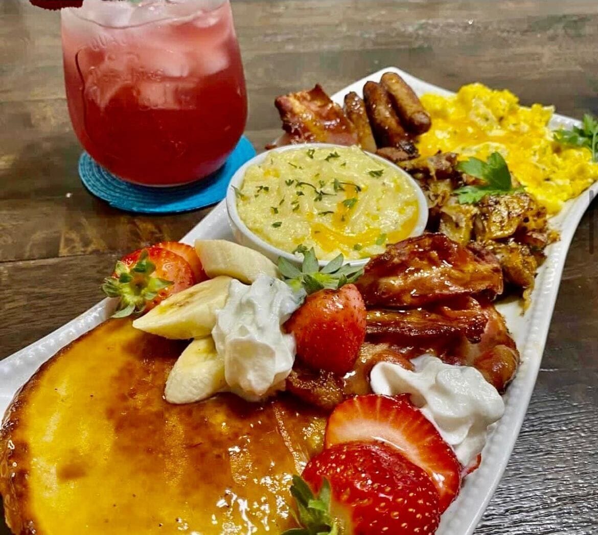 A breakfast plate with scrambled eggs, bacon, toast, strawberries, bananas, whipped cream, and a drink.