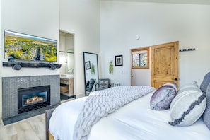 Un dormitorio con chimenea, cama con almohadas, televisor y puerta con espejo y cuadros.