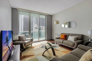 Un salon moderne comprenant un canapé, des fauteuils, une table basse en verre, un téléviseur à écran plat et une grande fenêtre donnant sur la ville.