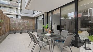 A modern outdoor patio with a dining table and chairs, surrounded by potted plants and a glass enclosure.