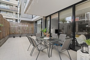 Une terrasse extérieure moderne, dotée d’une table et de chaises, entourée de plantes en pot et protégée par une paroi en verre.