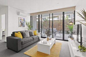 Un salon moderne avec un canapé gris, une table basse blanche et une grande fenêtre donnant sur un jardin et des immeubles.