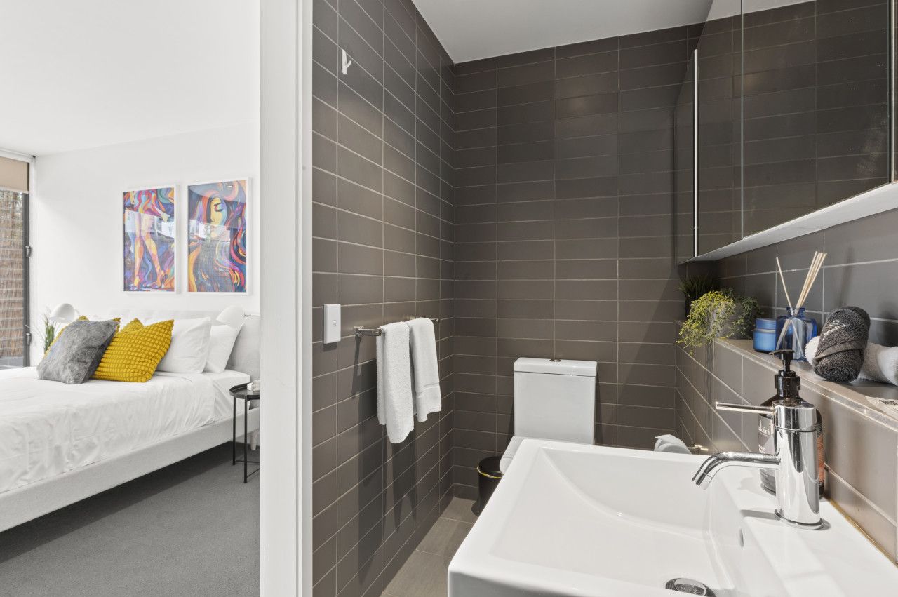 A modern bathroom with a white sink, a toilet, and a shower area. There is a bed with a white headboard and pillows in the adjacent room.