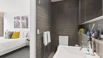 A modern bathroom with a white sink, a toilet, and a shower area. There is a bed with a white headboard and pillows in the adjacent room.
