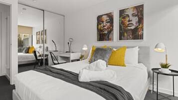 A modern hotel room with a large bed, a desk, and a chair. There are two paintings on the wall and a small table with a plant.