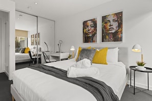 Une chambre d’hôtel moderne avec un grand lit, un bureau et une chaise. Il y a deux tableaux au mur et une petite table avec une plante.