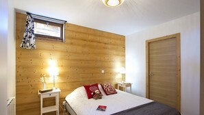 A bedroom with a wooden headboard, two bedside lamps, a bed with red and patterned pillows, a small bedside table, and a window with a curtain.