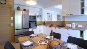 A modern kitchen with a built-in oven, microwave, and refrigerator. There is a dining table with chairs, a clock on the wall, and a hanging light fixture.