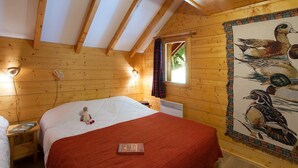 A cozy wooden cabin bedroom with a bed, a nightstand, a reading lamp, and a window with a view of greenery.