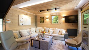 A cozy living room with a wooden ceiling, a large sofa, a coffee table, and a TV mounted on the wall.