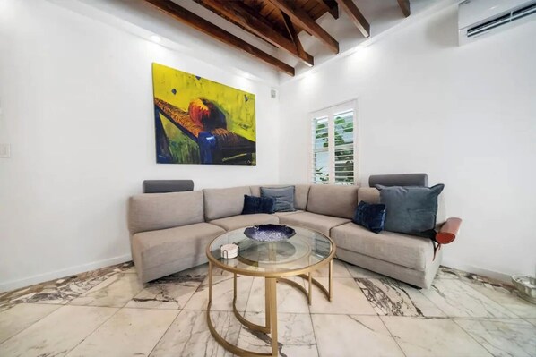 Ein Wohnzimmer mit einem beigefarbenen Ecksofa, einem Glastisch und einem farbenfrohen abstrakten Gemälde an der Wand.
