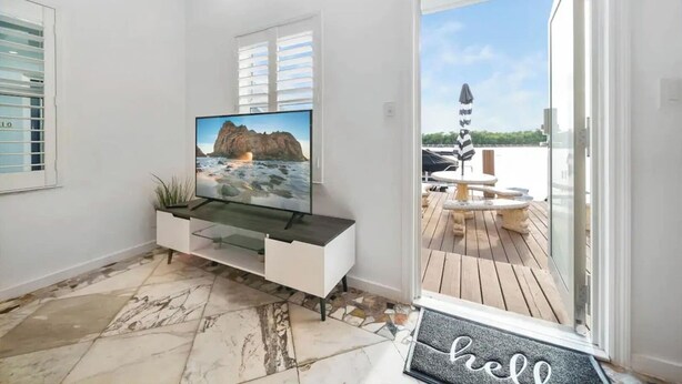 A modern living room with a large flat-screen TV on a glass-topped console, a doormat with "hello" on it, and a view of a wooden deck and water outside. - Waterside Escape w/Queen Bed and Free Parking (Hollywood)