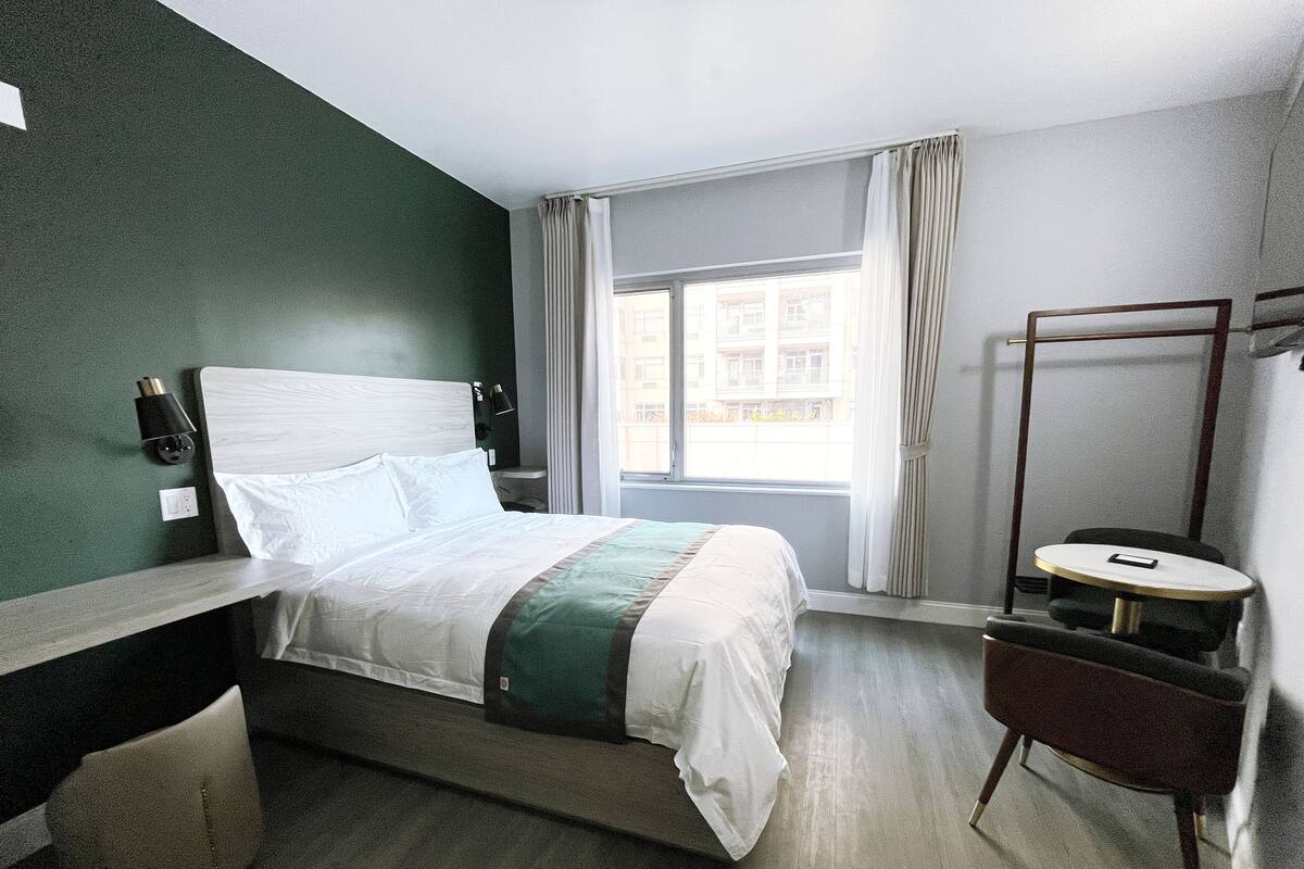 Hotellihuone, jossa on sänky, yöpöytä, tuoli ja ikkuna, jossa on verhot.